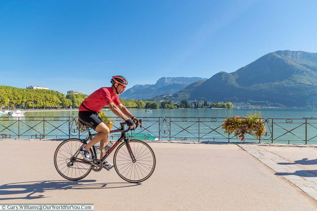 A road cyclist, in bright red, cycling around Lake Annecy, with the green mountains as a backdrop
