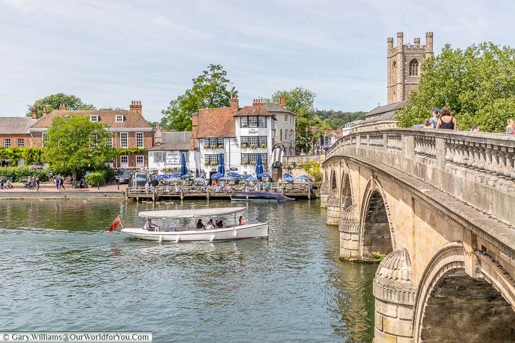 A small pleasure boat heading under the Henley Bridge in front of the The Angel on the Bridge pub in Henley on Thames