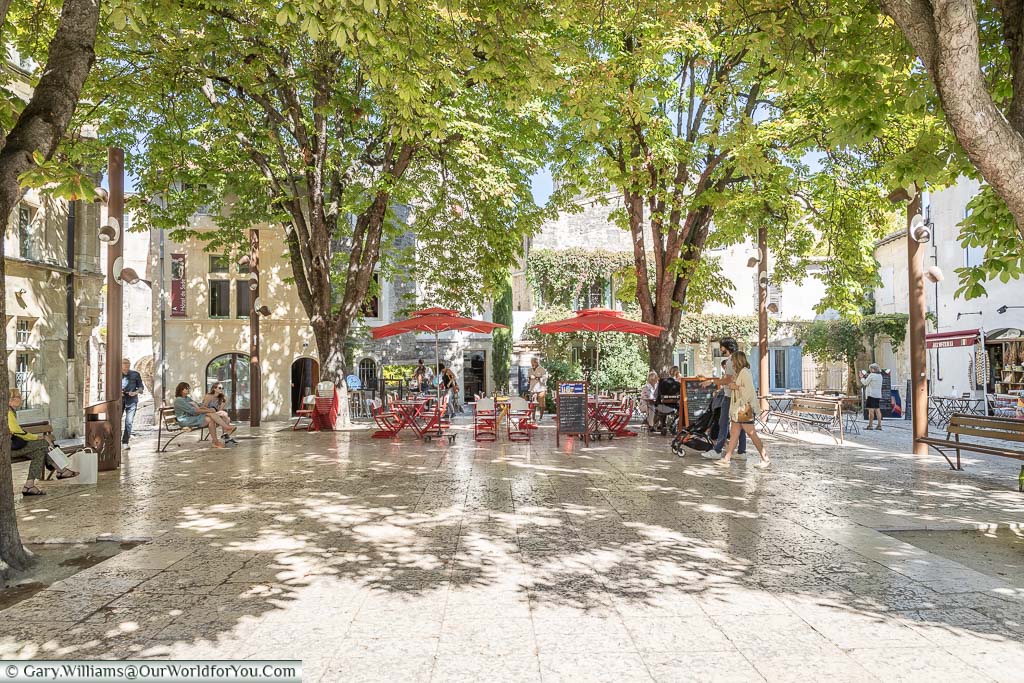 A bright square, called Place Favier, in Saint-Rémy-de-Provence under the dappled shade with a couple of colourful red tables and chairs, under parasols, of a local restaurant.
