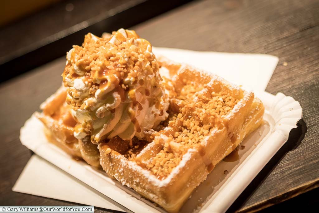 A crisp freshly cooked waffle topped with cream, caramel sauce and crunchy nuts all dusted with icing sugar in a paper plate on a German Christmas Market