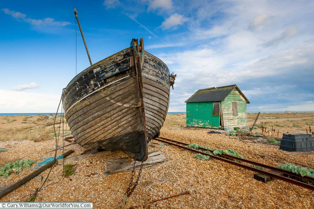 An abandoned fisherman's wooden trawler and dilapidated, green, wooden shack next to small track railway lines on the beach of Dungeness.