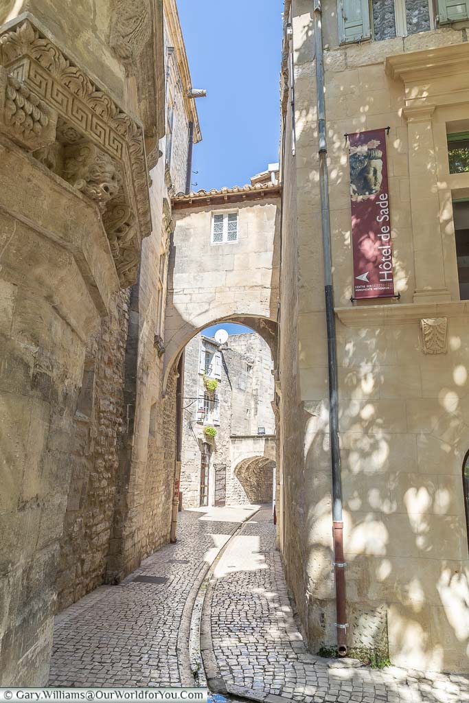 Looking a long and narrow cobbled passageway, under an archway, in centre of Saint Remy de Provence. A channel runs down the centre of the lane to allow rainwater to flow away.