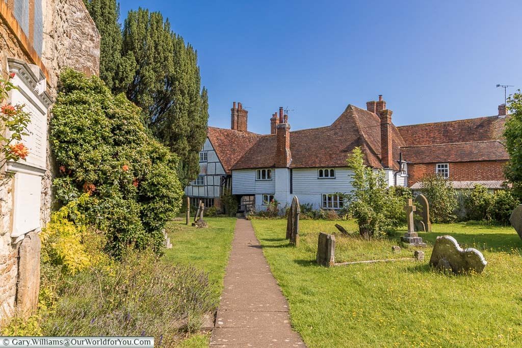 A view from the pathway in the churchyard of St Michael the Archangel looking towards white weather boarded buildings with red tiled roofs in Smarden, Kent