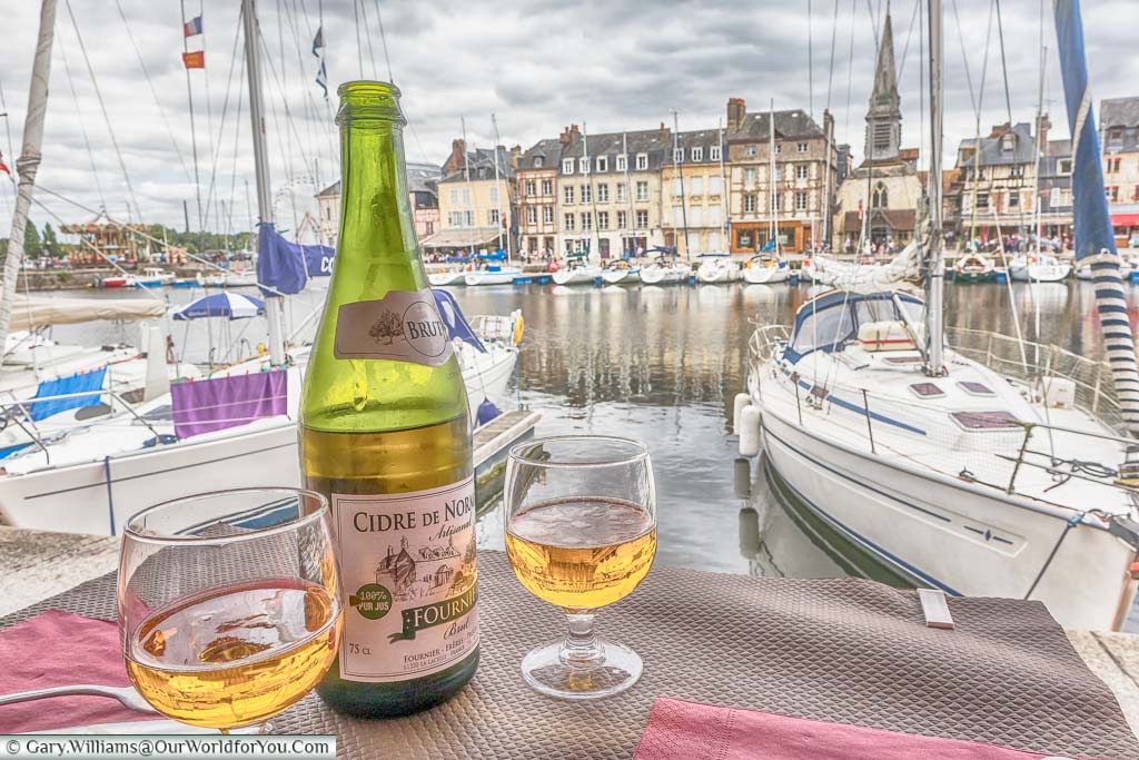A bottle and 2 glasses of Cidre de Normandie served at the edge of the marina of Honfleur, filled with small sailing boats.