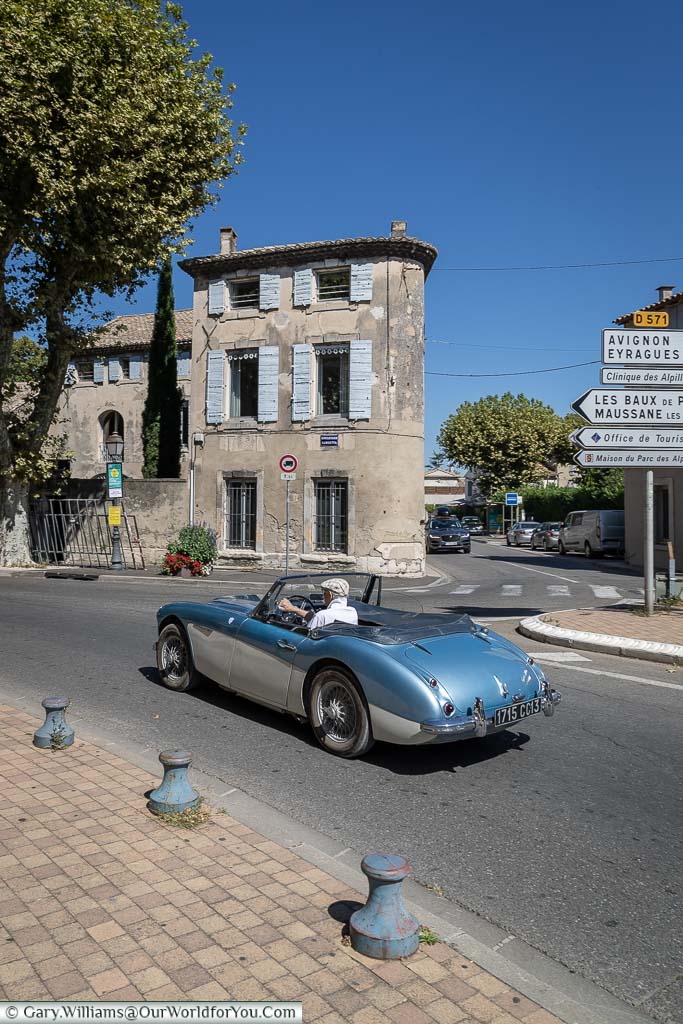 A classic 1960's Austin Healey convertible sports car in metallic blue & cream as it passes Saint-Rémy-de-Provence in front of a blue shuttered home on a beautiful sunny day.