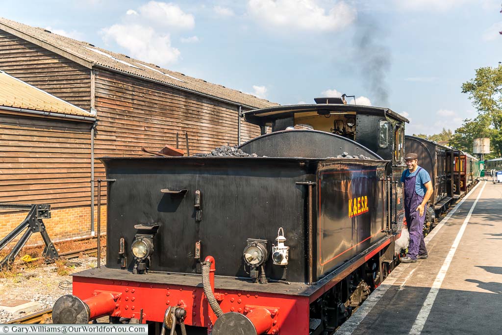 The back of a steam train with dark smoke coming from its chimney and the driver preparing to get on his way on a bright sunny day