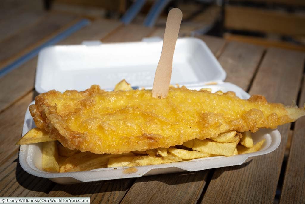 A battered golden fillet of cod, speared with a wooden fork, rested on a portion of chips in a polystyrene container from Great Yarmouth, Norfolk