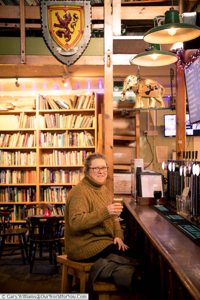 Janis wrapped up warmly for winter holding her beer inside the Chapel. The beer pumps are on the right hand side and a bookcase full of books behind her.