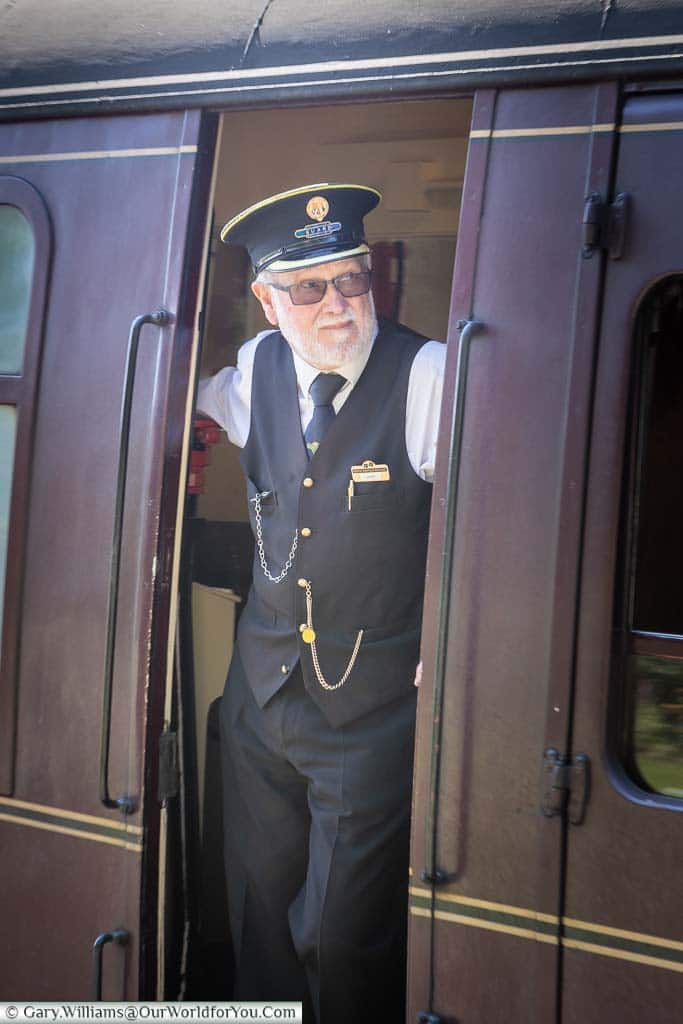 A well-dressed guard is standing in the doorway of the guards' carriage as a steam train prepares to depart the Sheringham station on the North Norfolk Railway.
