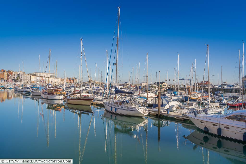 Small sailing boats in Ramsgate’s Marina resting on perfectly still water under a bright blue sky.