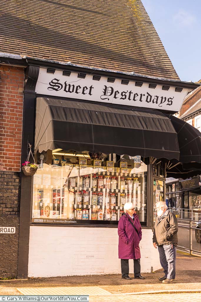 An elderly couple standing outside Sweet Yesterdays’ a traditional confectionery store with windows full of glass jars of different treats.