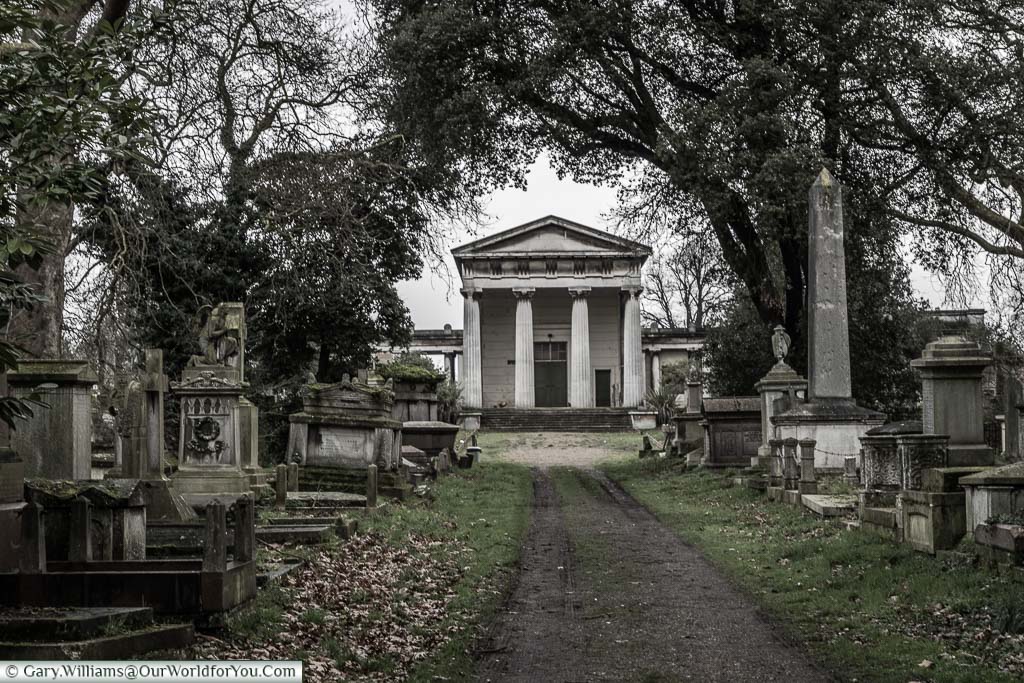 A path through Kensal Green Cemetery leading to the disused Anglican Chapel built in a neo-classical design with 4 Doric columns at the entrance. Either side of the way are tombs & gravestones.