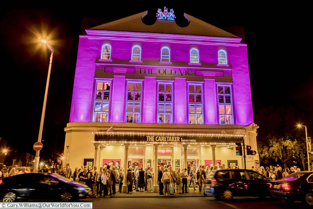 Crowds exiting the Old Vic Theatre at night. The foyer is illuminated in golden light and the top of the theatre is floodlit in a cerise pink