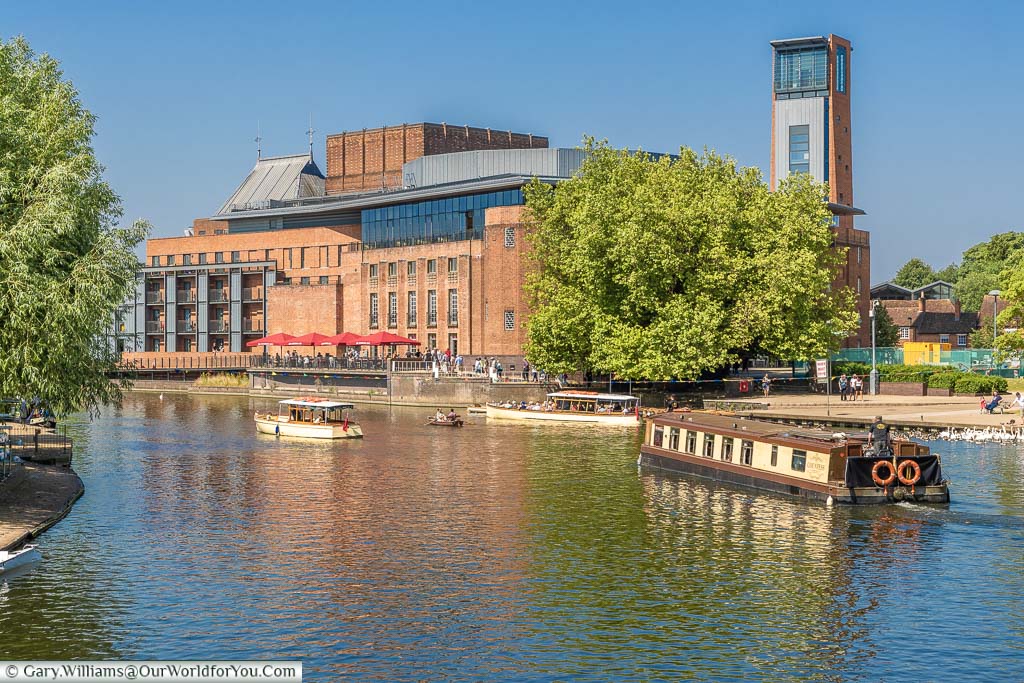 A wide-beam cnalboat navigating on the River Avon between other boats in front of the Royal Shakespeare Theatre on a beautiful sunny summers day.