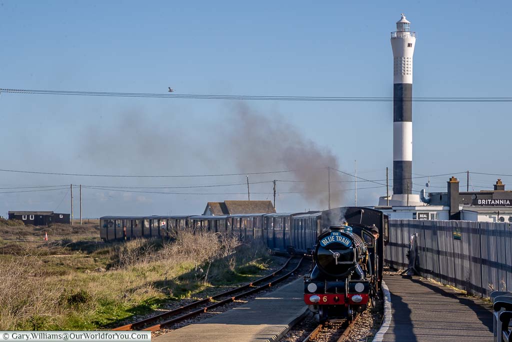 The modern, fifth, tall, Dungeness lighthouse in the background, with the Romney, Hythe and Dymchurch steam train, arriving at Dungeness station in the foreground.