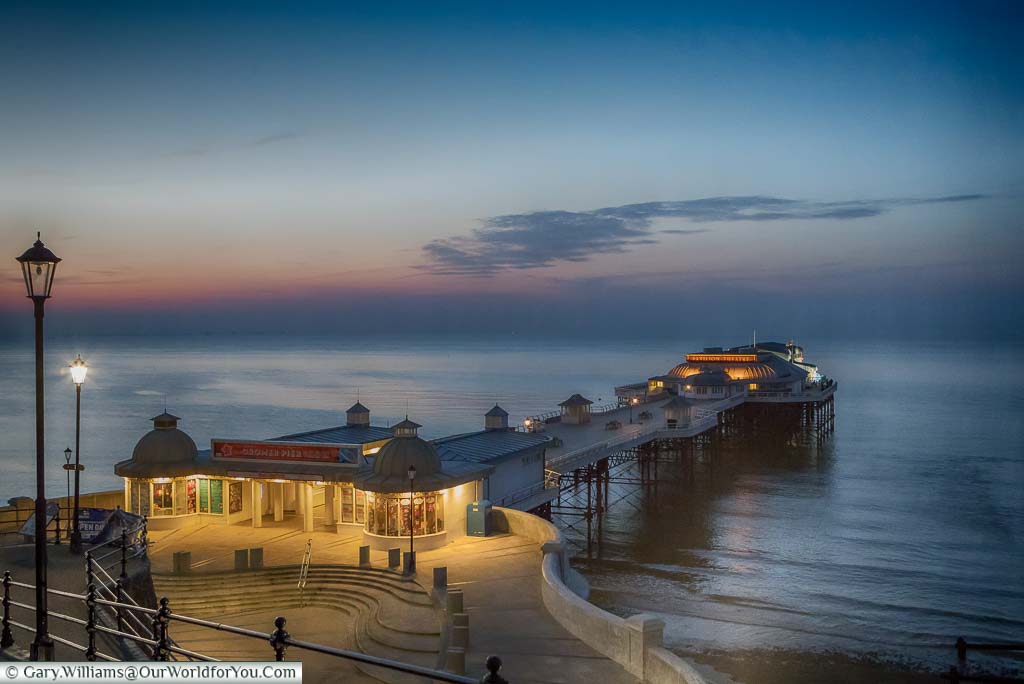 A view of the illuminated, elegant, Cromer Pier, after dusk, under orange and blue hues.
