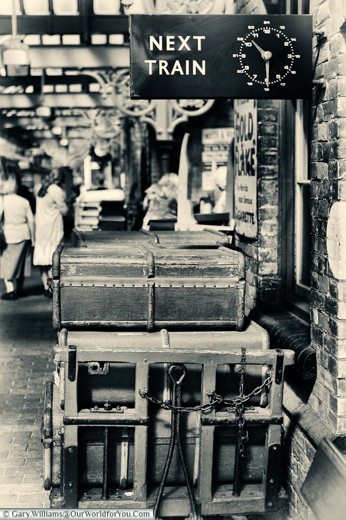 A nostalgic black and white image of a stack of traditional suitcases under a signed for the next train at the Sheringham station of the North Norfolk Railway.