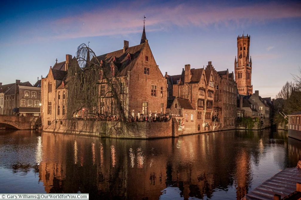 A view, at dusk, of the canals, and red brick medieval building of Bruges, with the Belfry in the backbround.