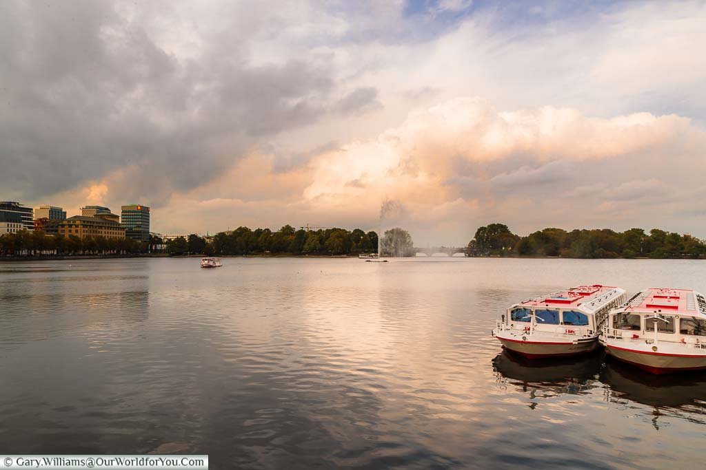 Looking across Binnenalster, a large man-made lake in the centre of Hamburg. There are Two pleasure boats moored up and the Alster Fountain in the distance.