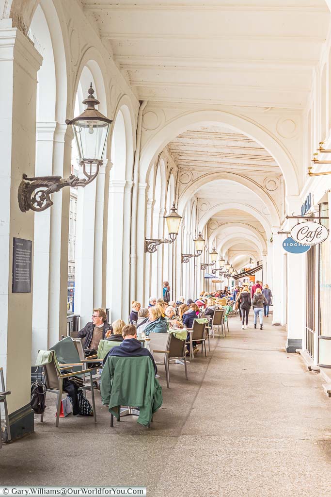 A look along the historic Venetian-style arcade along the waterfront of Hamburg with tables and chairs filled by cafe patrons.