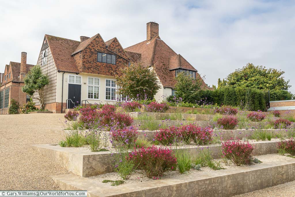 Raised flower beds with cerise coloured flowers leading to a pretty house at the entrance to RHS Gardens Wisley