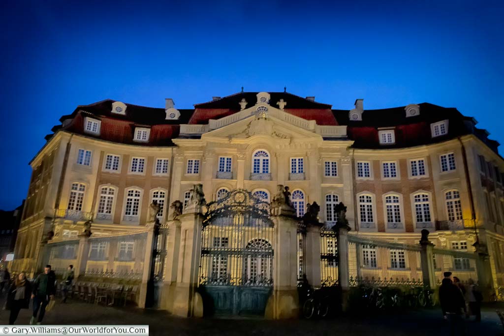 The Baroque Erbdrostenhof palace at dusk under a blue sky. The lit sandstone building stands imposingly over Salzstraße and Ringoldsgasse.