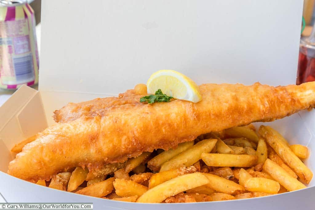 A portion of cod in crispy batter sitting on a bed of crispy garlic fries from the Smokehouse served in white cardboard packaging.