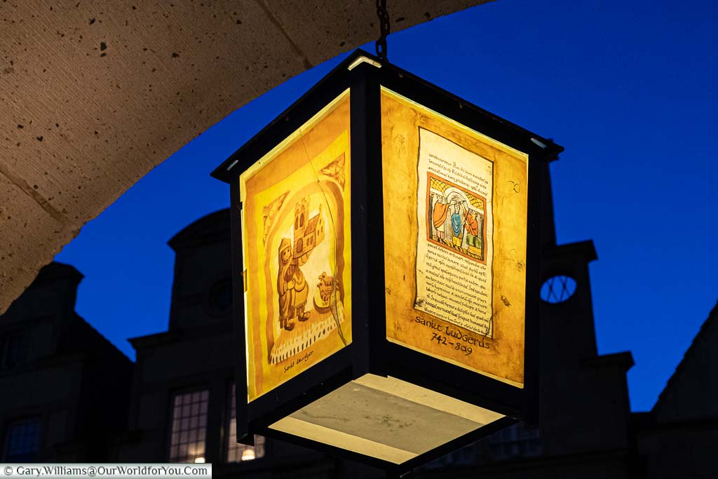 A lit orange lantern, handing from an arch in Prinzipalmarkt, depicting scenes from historic German tales against the deep blue of dusk and the rooftops of the merchants' buildings.
