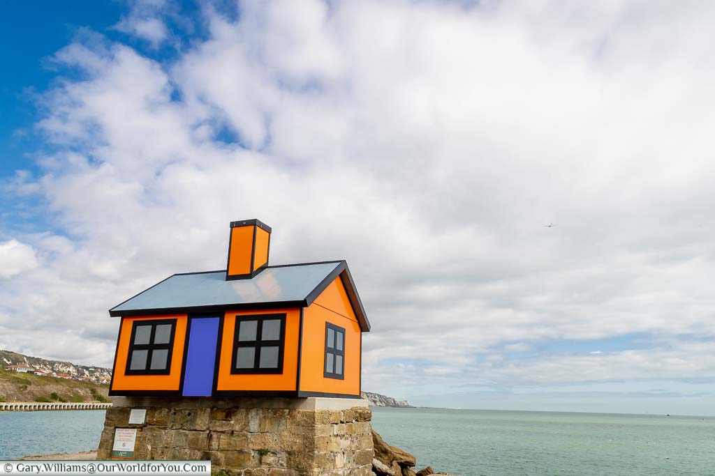 Holiday Home – By Richard Woods; An orange bungalow art installation with a purple door sitting on a plinth at the edge of the harbour car park overlooking the sea.