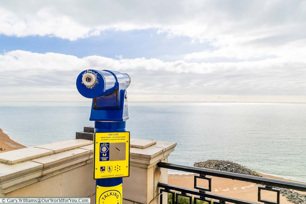 A blue fixed telescope overlooking the English change towards France as blue skies start to appear on an otherwise cloudy day.