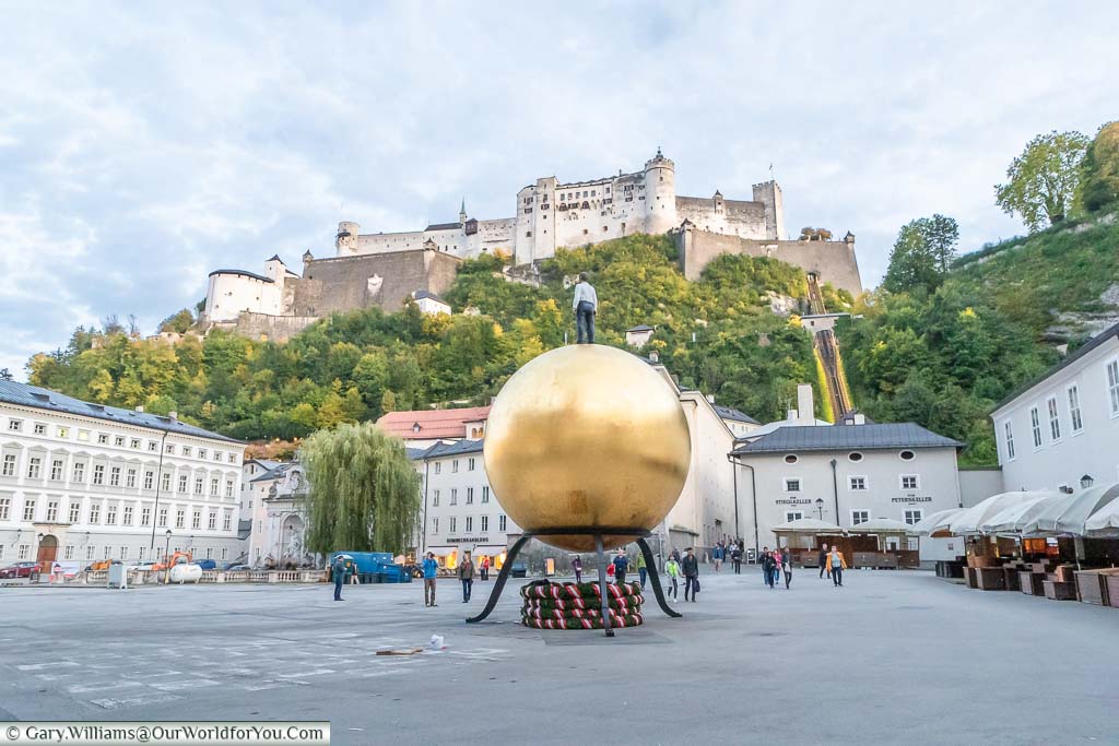 The modern art peice 'Sphaera', a figure of a man standing on a large golden orb in Kapitelplatz overlooked by Salzburg Castle in the early evening.