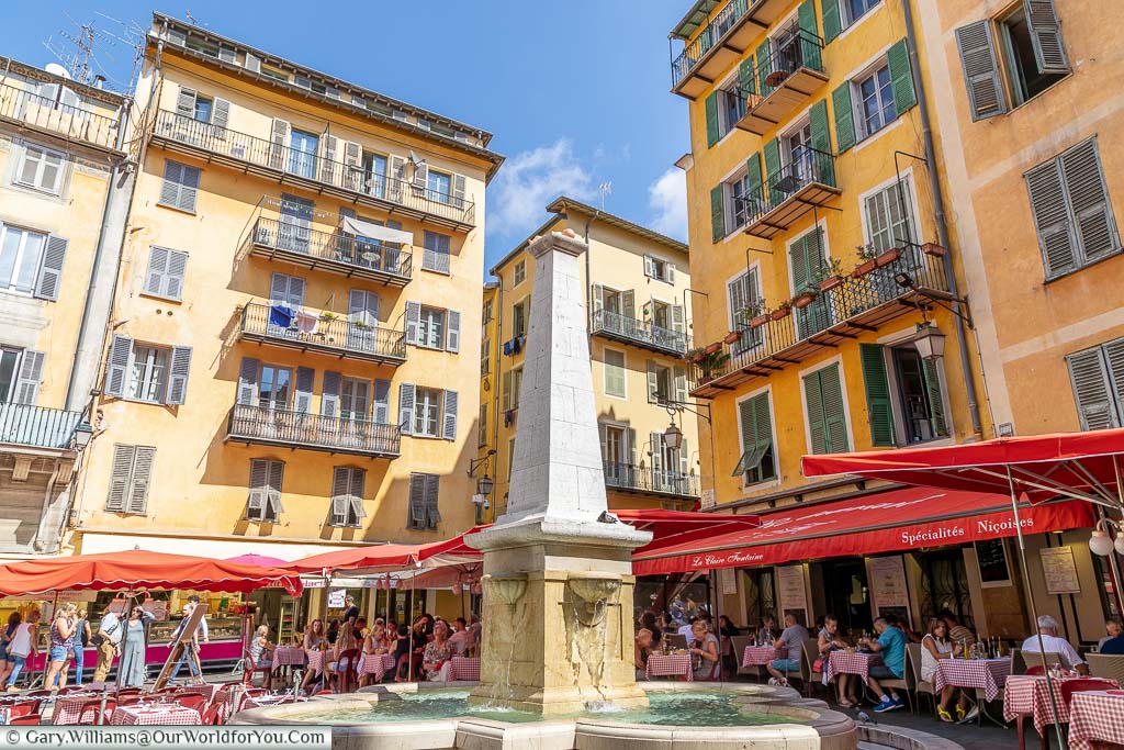 A fountain in a square in central Nice, surrounded by tables and chairs covered by parasols, Flanked on all sides by brightly coloured residential buildings.