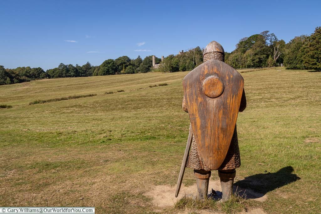 The view of the back of a carved wooden statue of a Norman Soldier overlooking the green rolling landscape of the battlefield at Battle with the Abby in the distance.