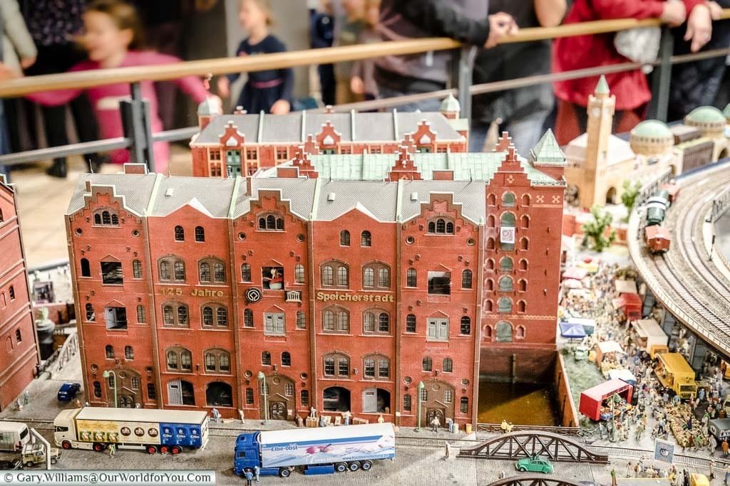 A model scene from Miniature Wonderland depicting the red brick warehouses of the Speicherstadt district with train tracks running alongside in front of the old Harbour building.