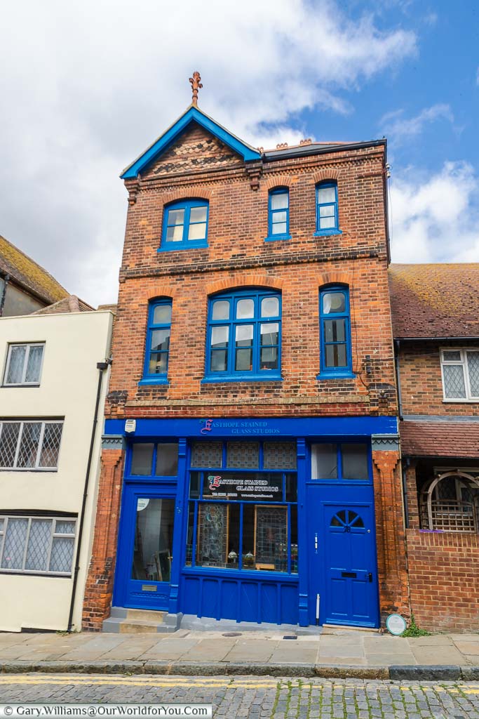A red brick three-storey building with a deep blue frontage, and blue window frames, that now houses a stained glass window shop.