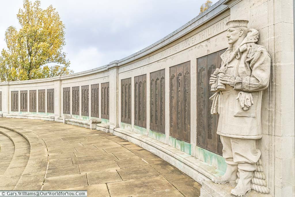 A stone seaman, holding binoculars, stands as a lookout at one end of a curved wall, adorned with brass plaques detailing the names & ranks of those lost at sea.
