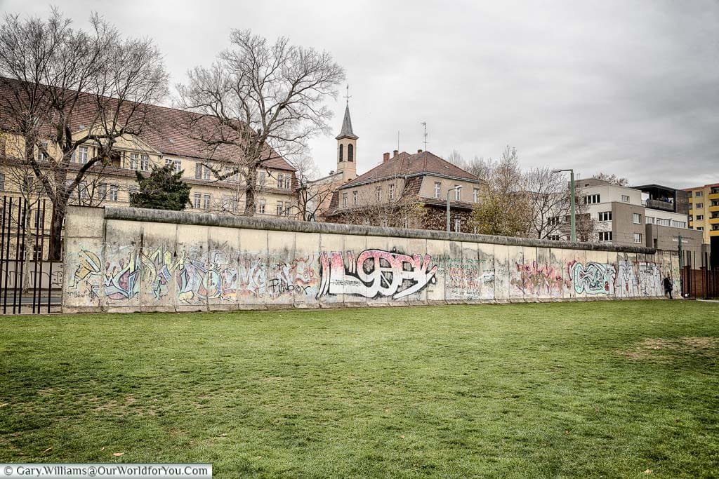 The graffitied Berlin Wall in front of a section of grass that made up the no-man's-land area in front of the wall on a grey December day.