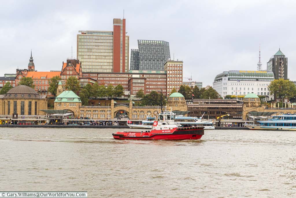 A red tug travelling along the Elbe River in front of the landing stages, against the backdrop of the St Pauli district of Hamburg.
