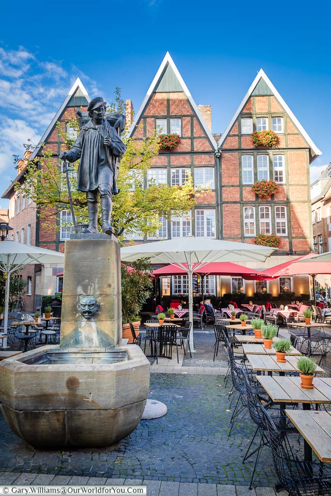A statue of a historical travelling merchant over a water fountain surrounded by tabes & chairs of the nearby reasturant, bars & cafes.