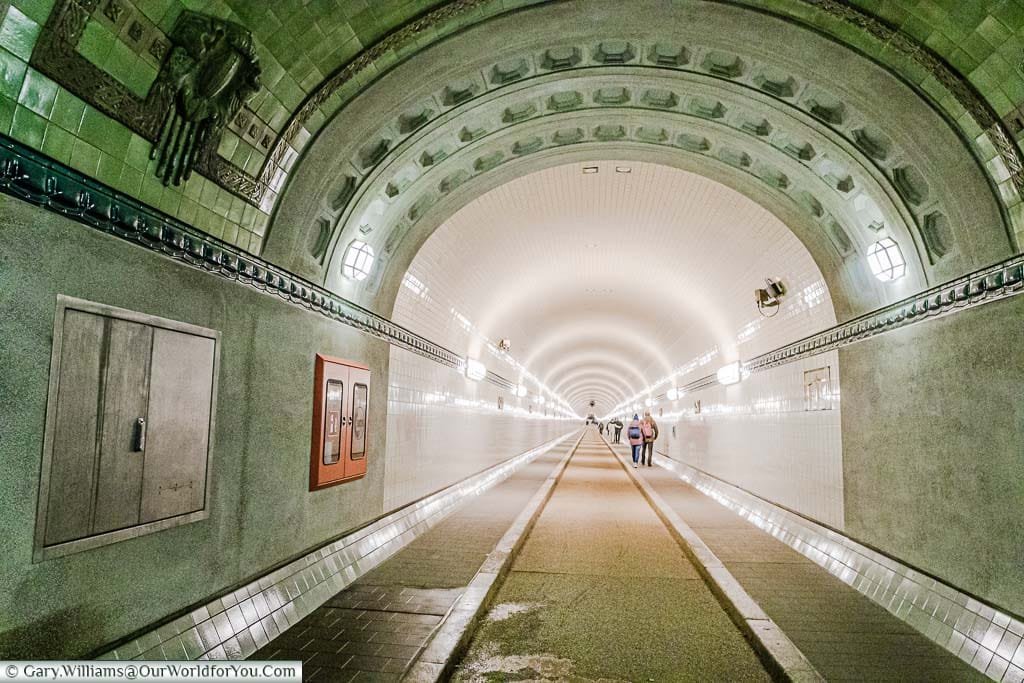 Looking along the recently restored, brightly lit, tiled tunnel That takes you under the Elbe River. There is a footpath on either side and a narrow track in the centre for cyclists that was once used by cars.