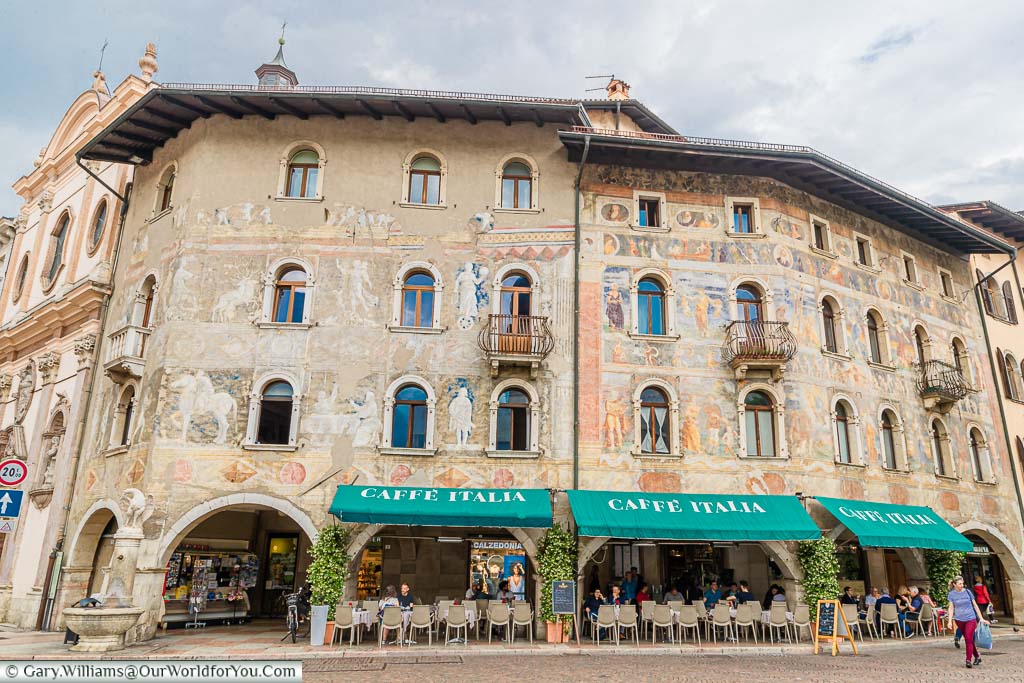 Café Italia in Piazza Duomo in the centre of Trento with green awnings covering the outdoor seating area with beautifully painted walls depicting classical Italian figures on the three storey renaissance building