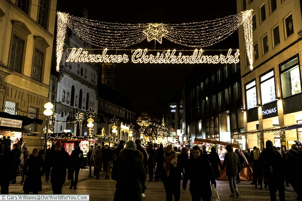 Pedestrians are walking along one of Munich shopping streets at night under fairy light telling you you are entering 'Müchner Christkindlmarkt.'