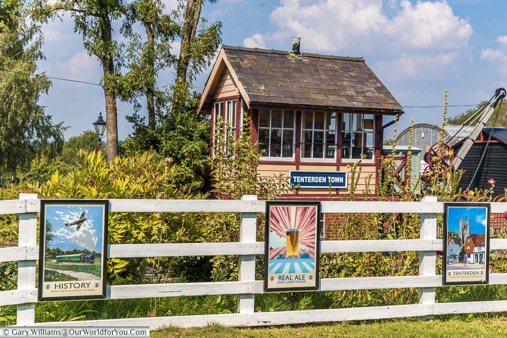 The signal box at Tenterden town steam railway with a white picket fence in front displaying posters of a bygone era.