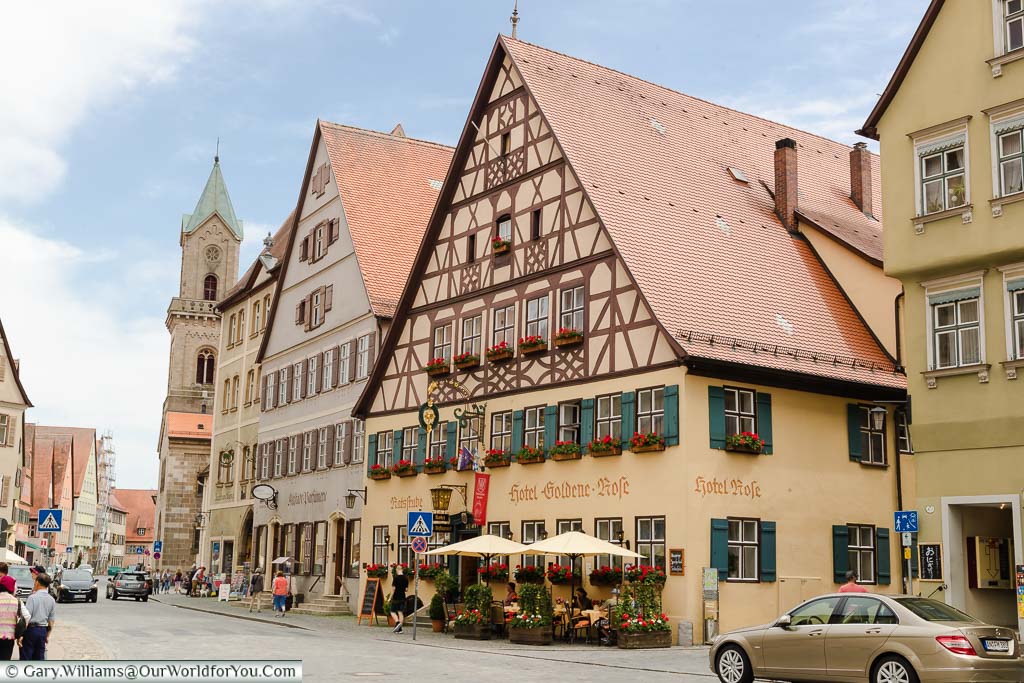 A street scene focused on the fabulous half-timbered Goldene Rose Hotel with window boxes full of red flowers with tables and chairs outside.