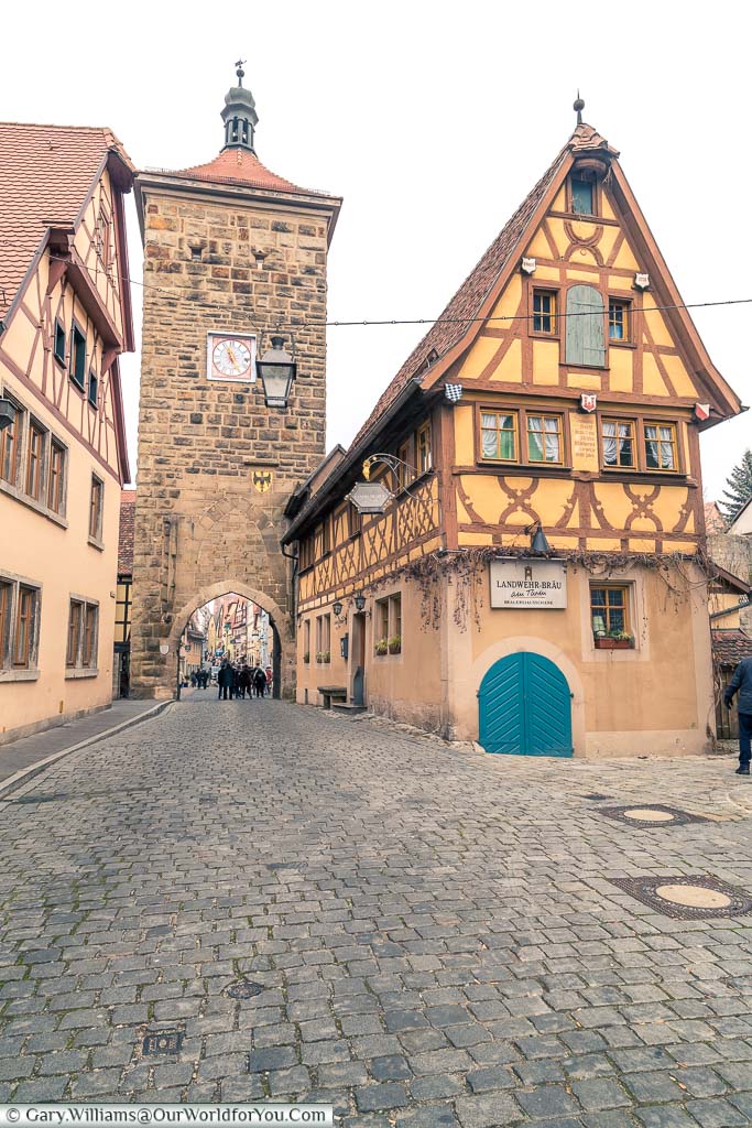 A view along one of the cobbled streets that lead into Rothenburg ob der Tauber through a gatehouse in the base of a tower. Alongside the tower is a typical, half-timbered, house with the steep roof of this area.