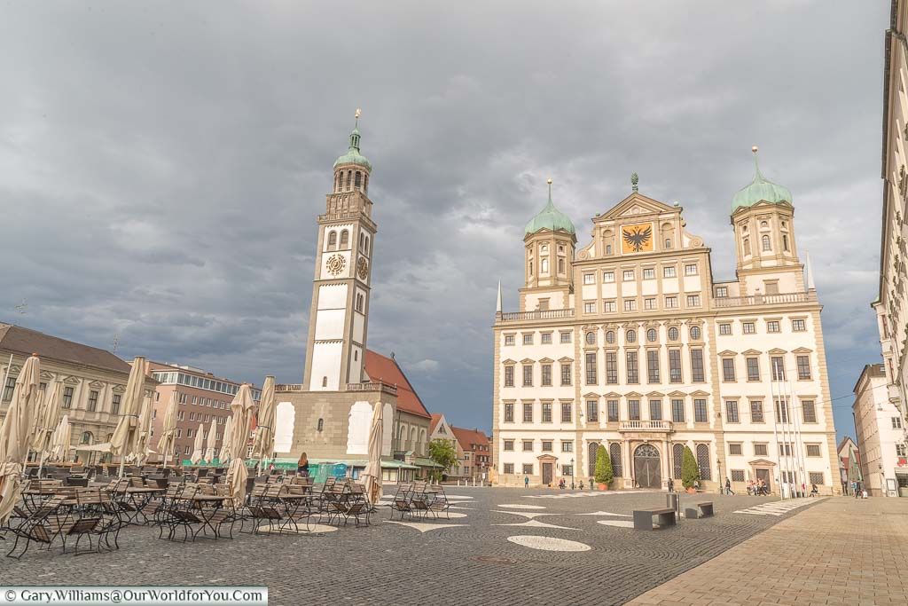 A view from the corner of Rathausplatz in Augsburg past a cafe's table and chairs towards the city's historic town hall. The Rathaus has two verdigris domes and sits alongside a church with a matching tower