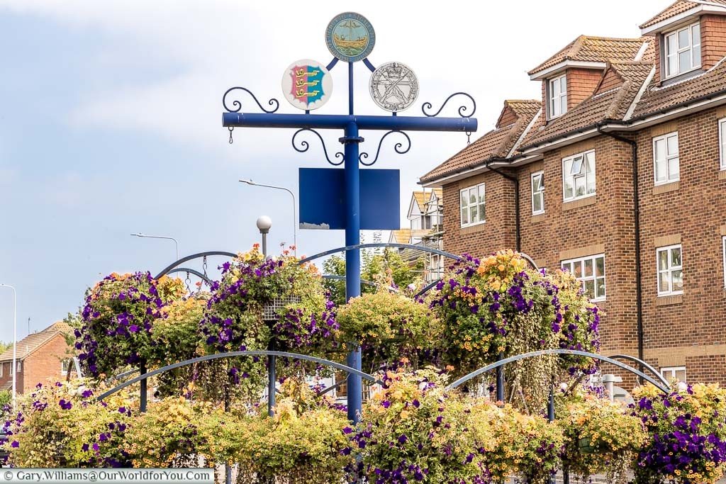 A roundabout decorated with flower baskets at the entrance to Hythe displaying signs for the town, its status as a Cinque Port and one from the Small Arms School