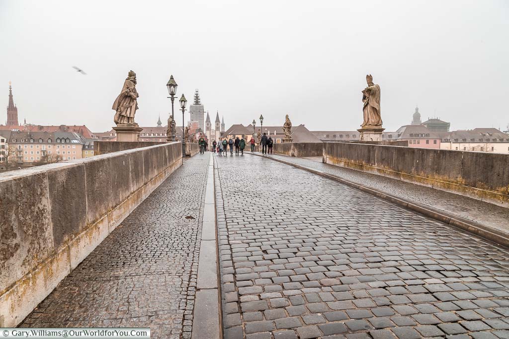 Strolling across the cobbled old main bridge in Würzburg on a damp, grey, day. The bridge is intersected on either side by stone statues of religious figures.