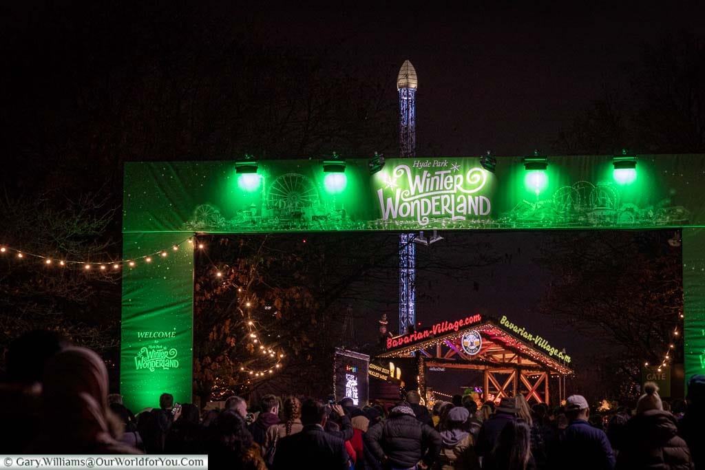 Crowds in front of the green entrance to Hyde park's Winter Wonderland with stalls from the barbarian village in the background.
