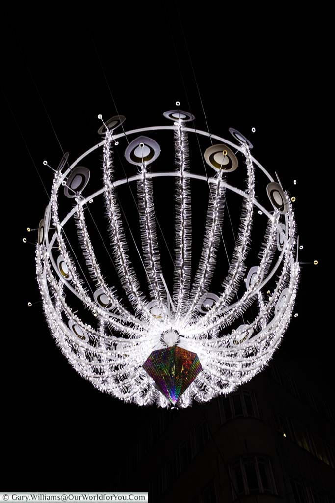 A close-up of an illuminated Christmas light feature hanging above New Bond Street in London’s West End. The ribs of the bauble resemble silver peacock feathers.